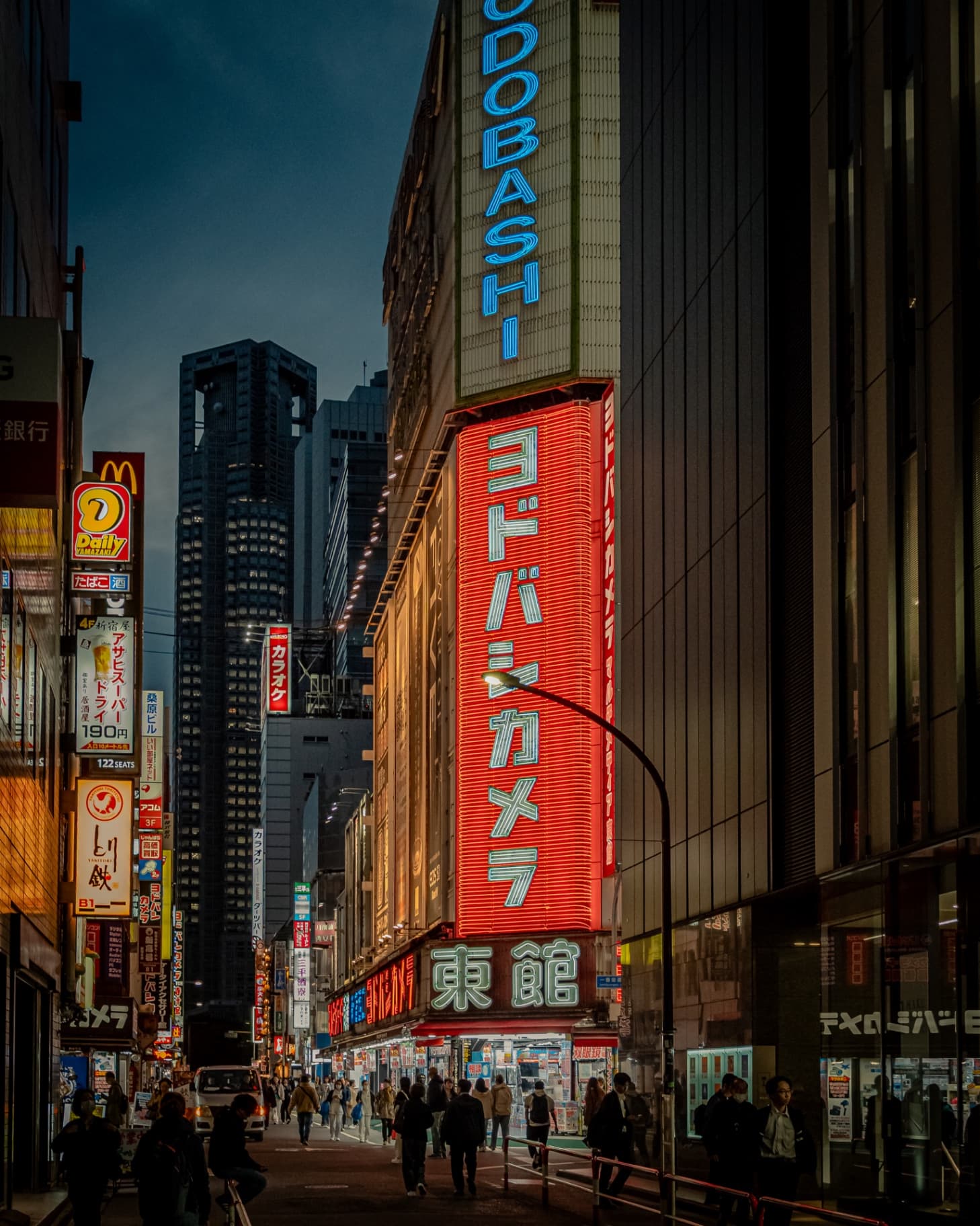 A busy Shinjuku street at night featuring a large red neon Yodobashi Camera sign with the Tokyo Metropolitan Government Building in the background.
