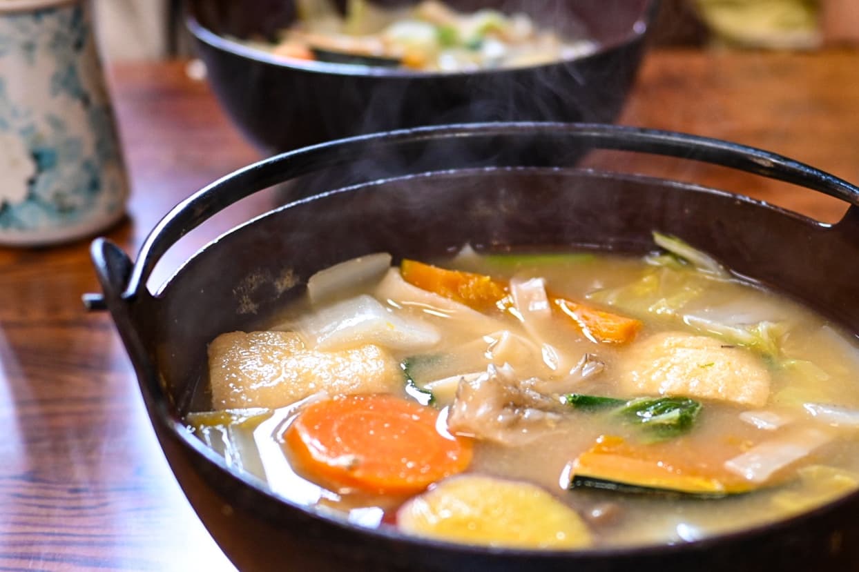 A close-up shot of a steaming iron pot containing Houtou noodles, pumpkin slices, carrots, and fried tofu in a rich miso soup.