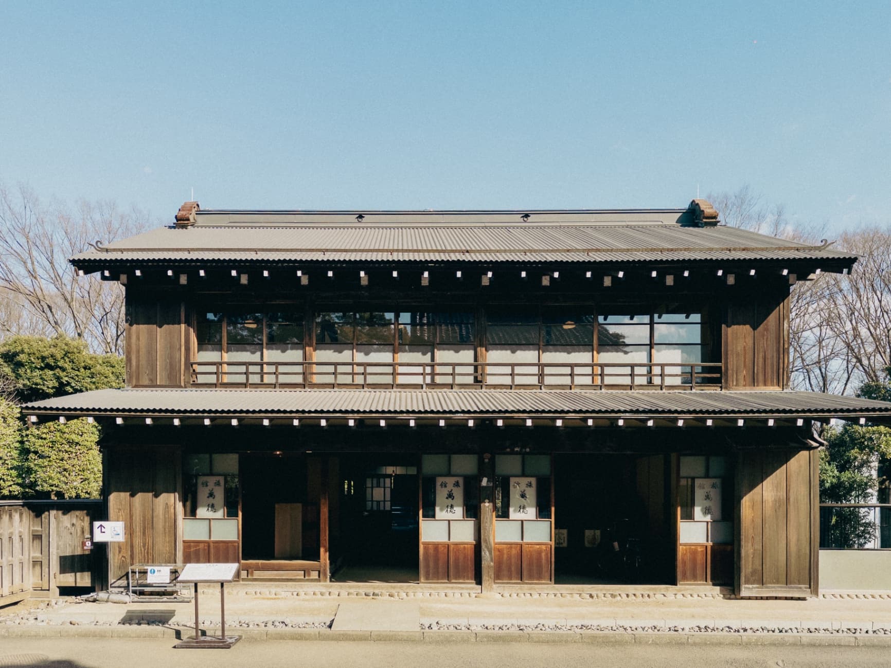 A two-story traditional Japanese wooden building with dark timber, sliding doors, and a tiled roof under a bright blue sky.