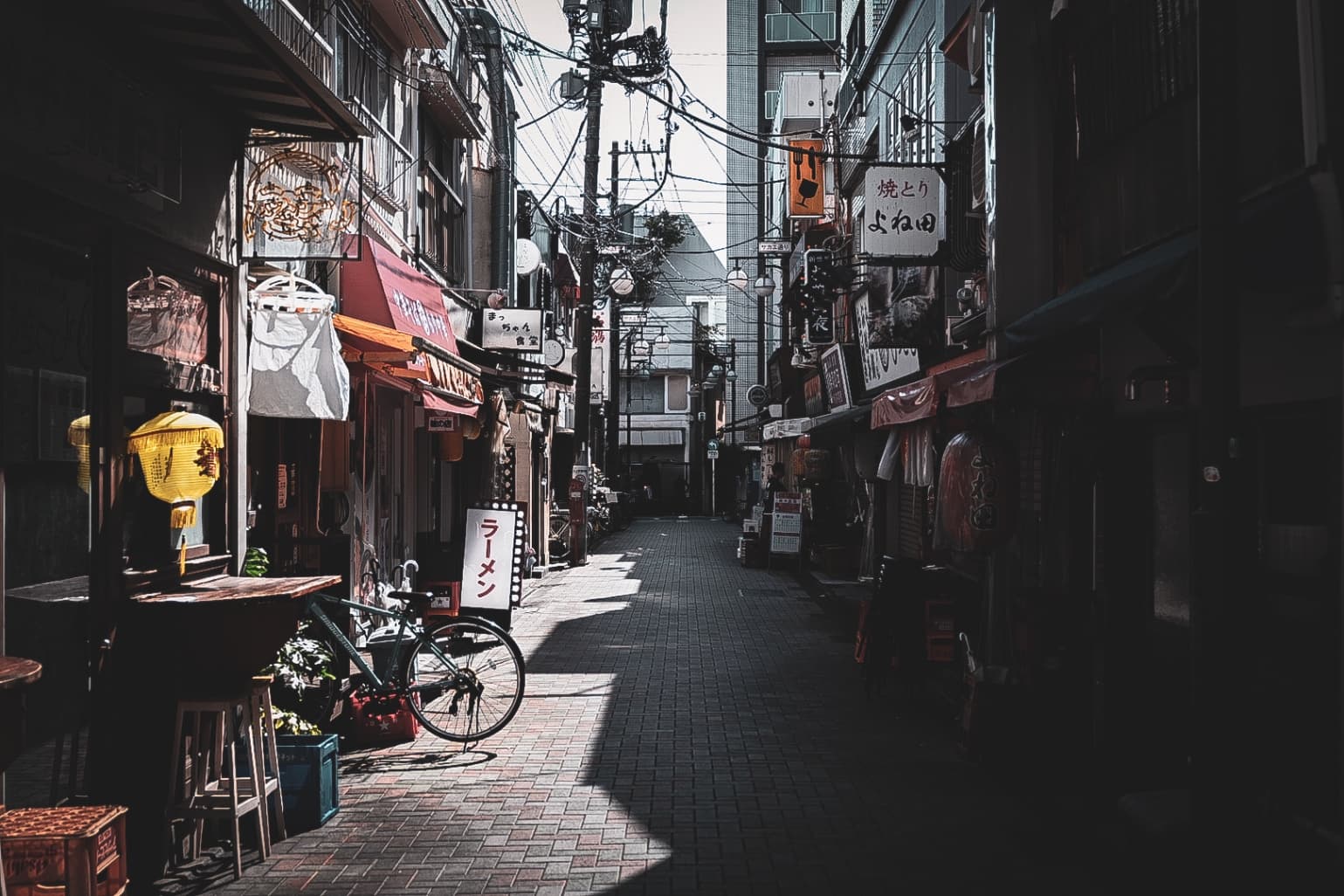 A narrow Japanese alleyway with restaurant signs, a bicycle, and power lines under bright sunlight.
