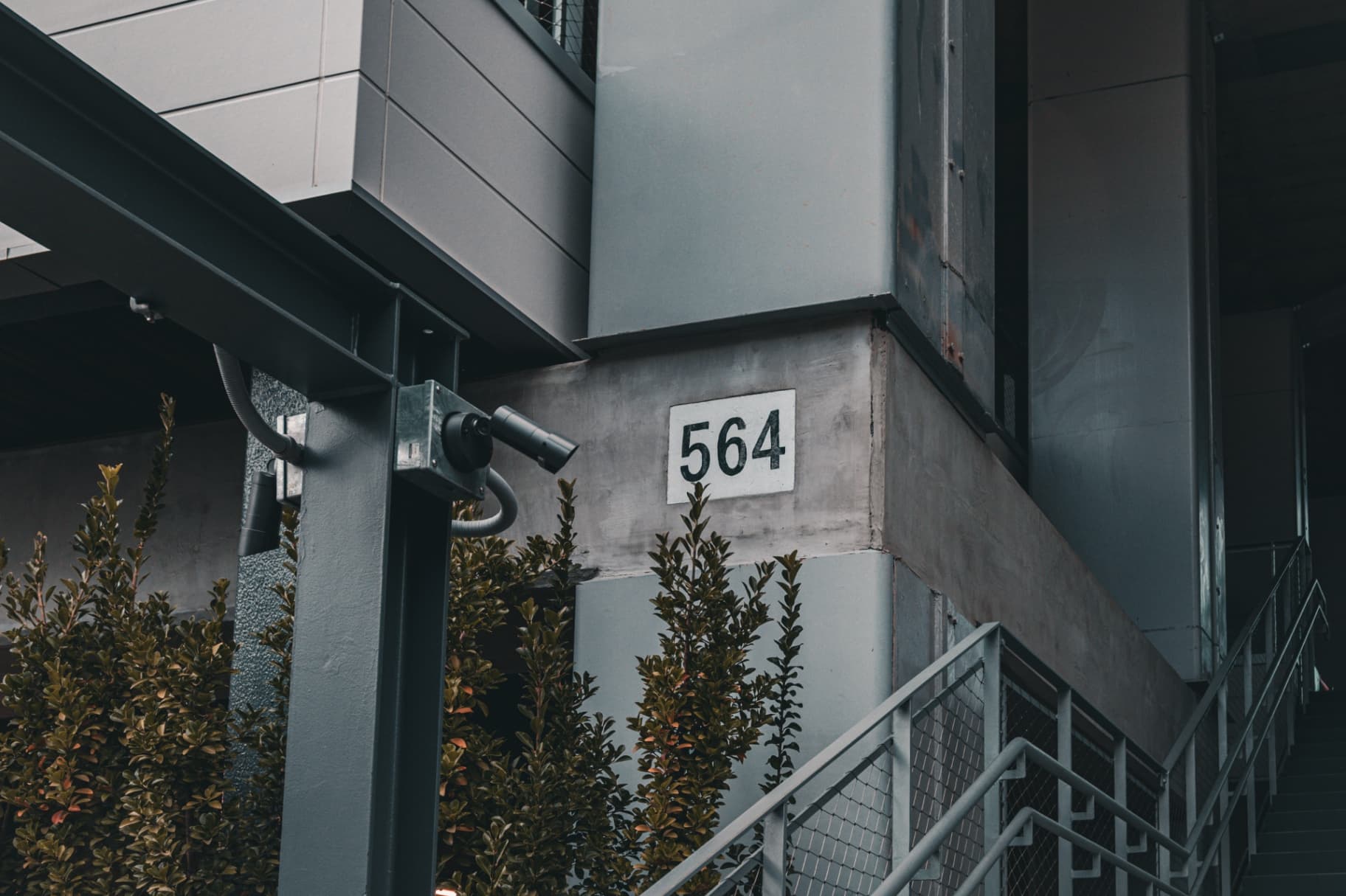 An industrial building exterior featuring the number 564 on a concrete wall with a security camera and green bushes in the foreground.