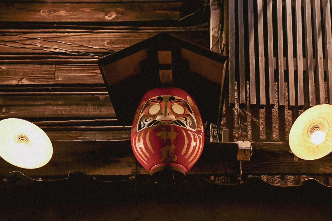 A red Daruma-shaped lantern hanging under a small roof between two round glowing lights against a wooden wall.