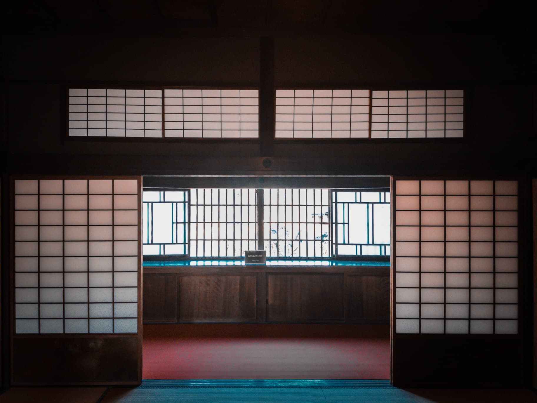A dark interior view through open shoji doors towards a bright latticed window in a traditional Japanese house.