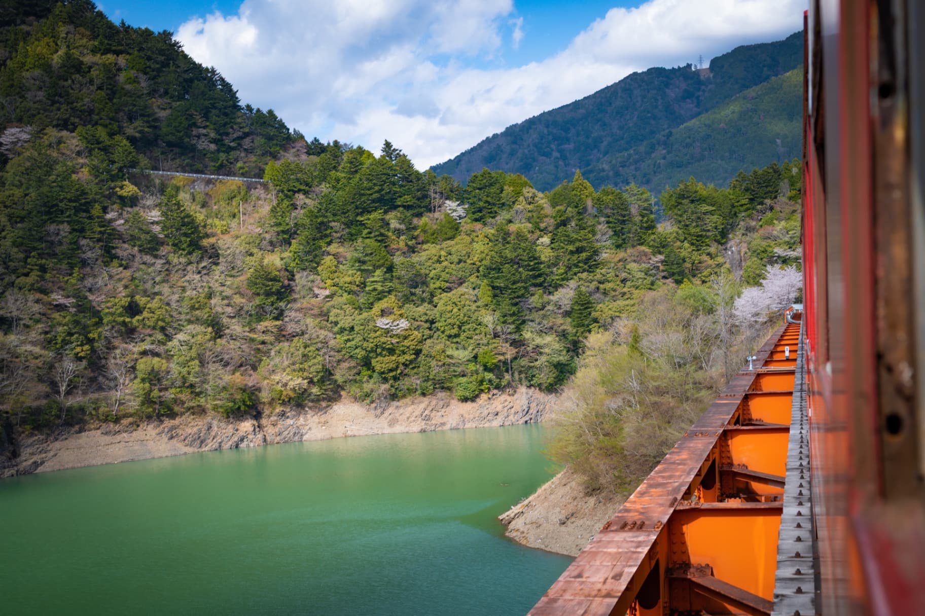 View from a train carriage crossing an orange steel bridge over a green lake with forested mountains in the background.