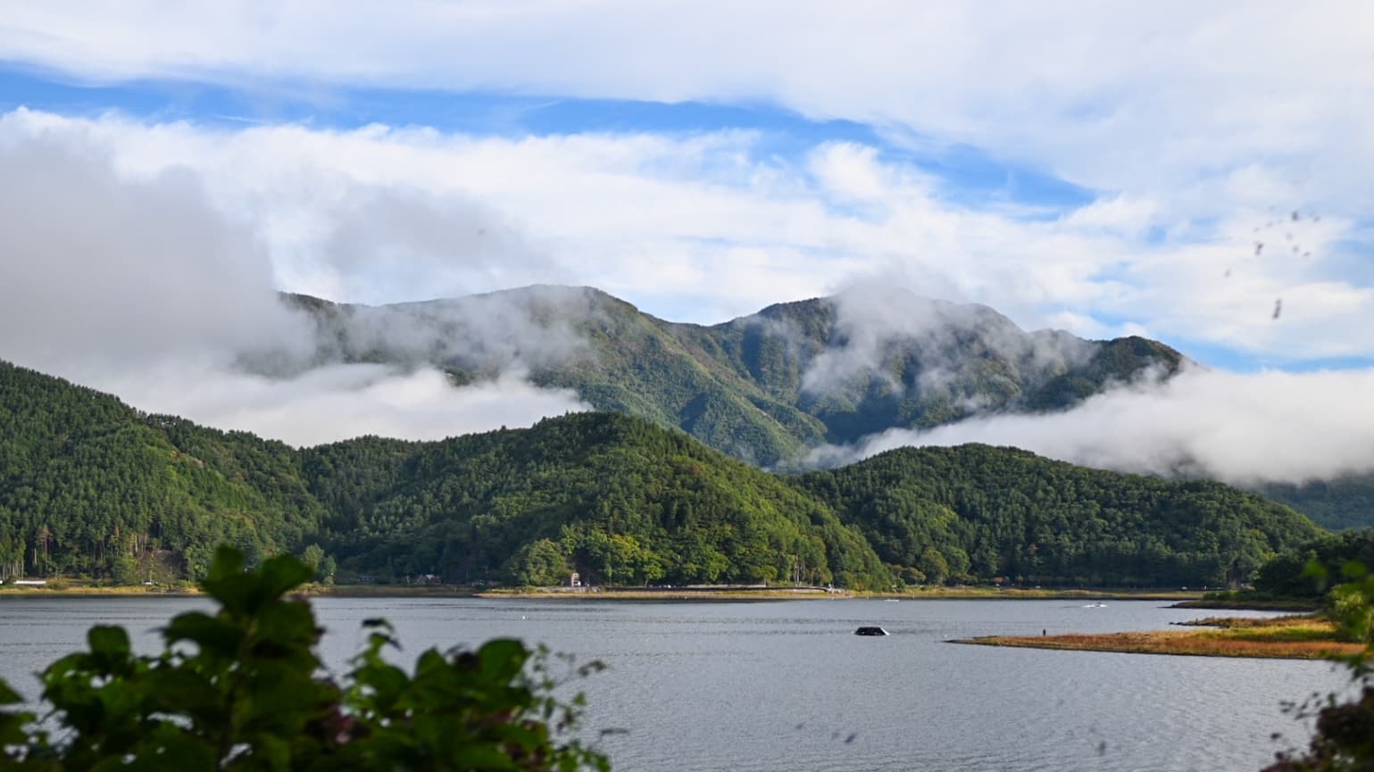A wide landscape view of Lake Saiko with misty green mountains and a blue sky.