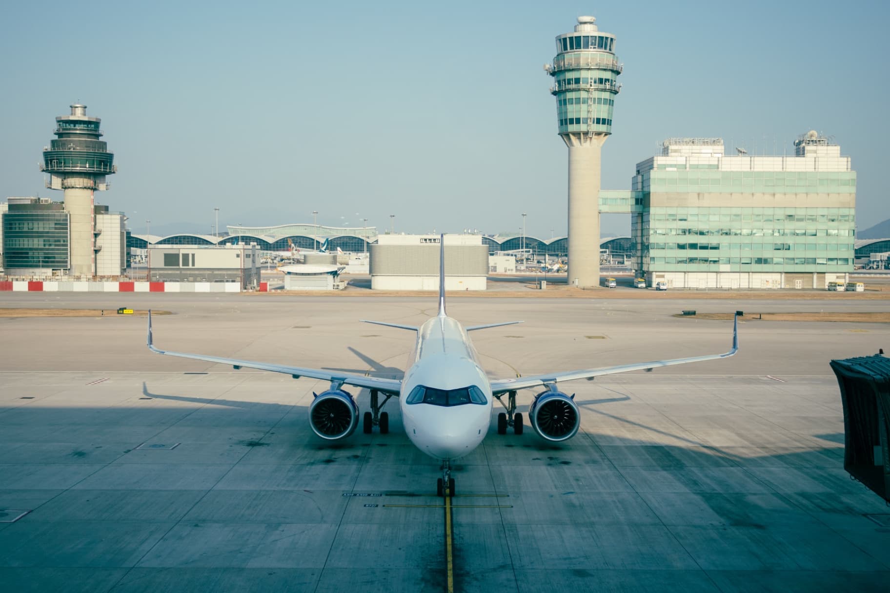 Front-on view of a white jet airplane at an airport gate with control towers and terminal buildings under a clear sky.