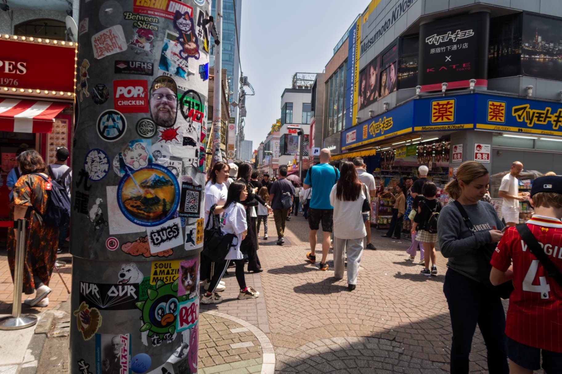 A utility pole in the foreground covered in various colorful stickers with a crowded shopping street and a Matsumoto Kiyoshi store in the background.